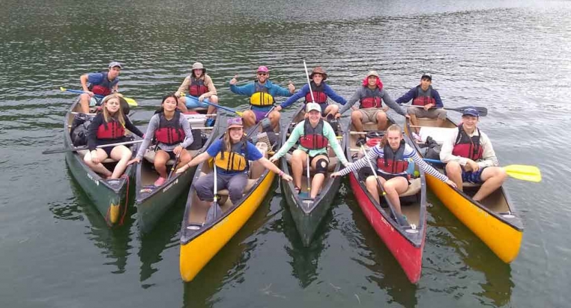 a group of students in six canoes smile for a photo on calm water